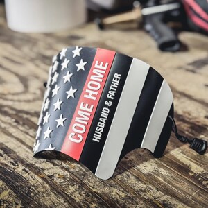 May include: A curved, semi-rigid black, white, and red accessory featuring a stylized American flag design. The text "COME HOME" is printed in white, followed by "HUSBAND & FATHER" in smaller white lettering. A black cord with a toggle is attached.