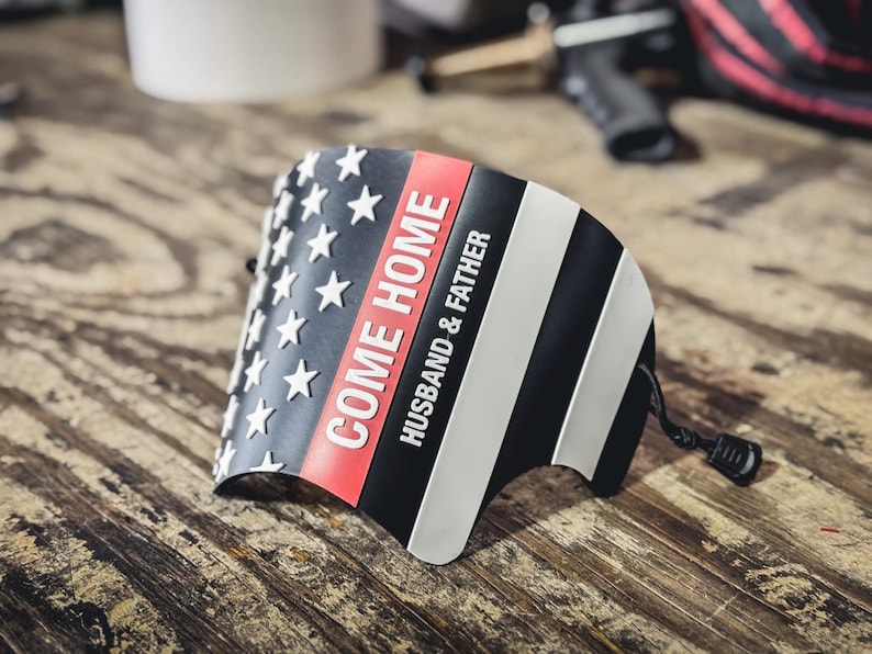 May include: A curved, semi-rigid black, white, and red accessory featuring a stylized American flag design. The text "COME HOME" is printed in white, followed by "HUSBAND & FATHER" in smaller white lettering. A black cord with a toggle is attached.