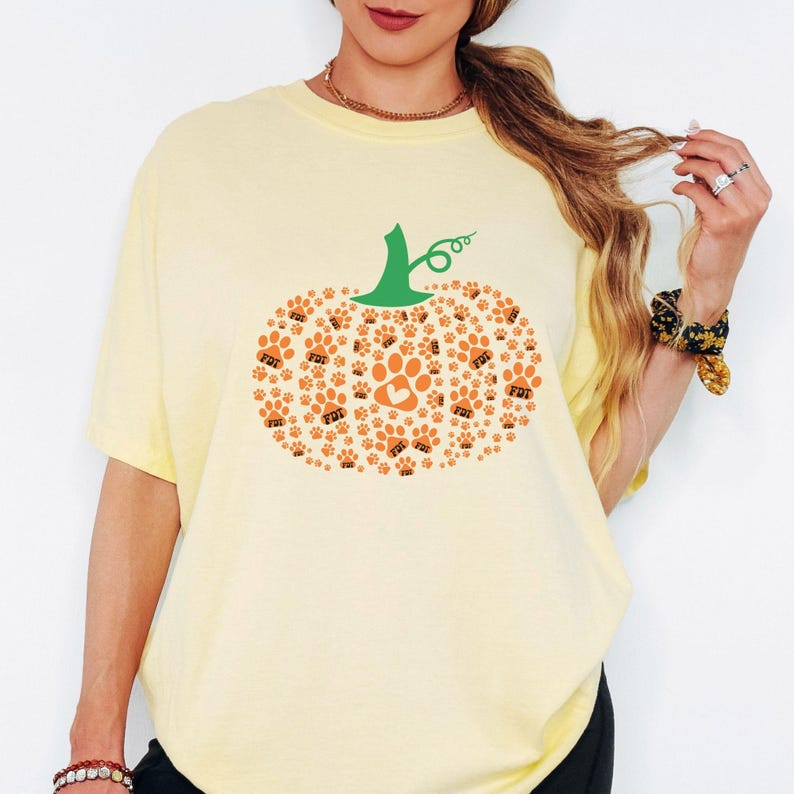 Puede incluir: Camiseta amarillo claro con un dise&ntilde;o de calabaza hecho de huellas de patas naranjas y un tallo verde. El dise&ntilde;o de la calabaza incluye una huella de pata con un coraz&oacute;n en el centro. La camiseta es de estilo c&oacute;modo e informal.
