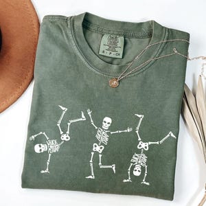 K&ouml;nnte beinhalten: Olivgr&uuml;nes T-Shirt mit wei&szlig;er Skelettgrafik. Das Shirt zeigt drei Skelette in spielerischen Posen. Eine goldene Halskette mit Anh&auml;nger ist &uuml;ber dem Shirt drapiert. Das Shirt ist mit "Comfort Colors" gekennzeichnet.