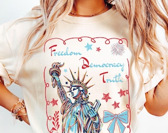 May include: A cream-coloured t-shirt featuring a graphic of the Statue of Liberty in blue and red tones. The text reads Freedom, Democracy, and Truth. The design includes red stars, a blue bow, and a firework, all enclosed in a red wavy border.