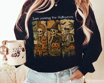 May include: A black long-sleeve sweatshirt featuring a graphic print with the text "Just Waiting for Halloween" above three panels showing skeletons in various activities, including wearing a towel, a witch hat, and sunglasses.