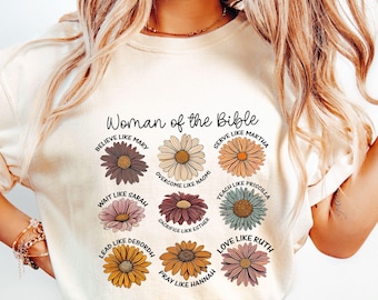 May include: Cream-colored t-shirt featuring the text "Woman of the Bible" above nine colorful daisy illustrations. Each daisy has a different color and a phrase such as "Believe like Mary" or "Love like Ruth".