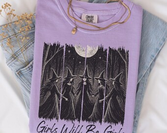 May include: A lilac-colored Comfort Colors t-shirt featuring a black and white graphic of five witches holding hands in a forest under a full moon. The text "Girls Will Be Girls" is printed below the graphic. A gold necklace rests on the shirt, which is folded on top of blue jeans.