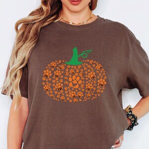 Puede incluir: Camiseta marr&oacute;n con un dise&ntilde;o de calabaza hecho de huellas de patas naranjas y un tallo verde. El dise&ntilde;o de la calabaza incluye una gran huella de pata en el centro. La camiseta tiene las letras "FDT" impresas en algunas de las huellas.