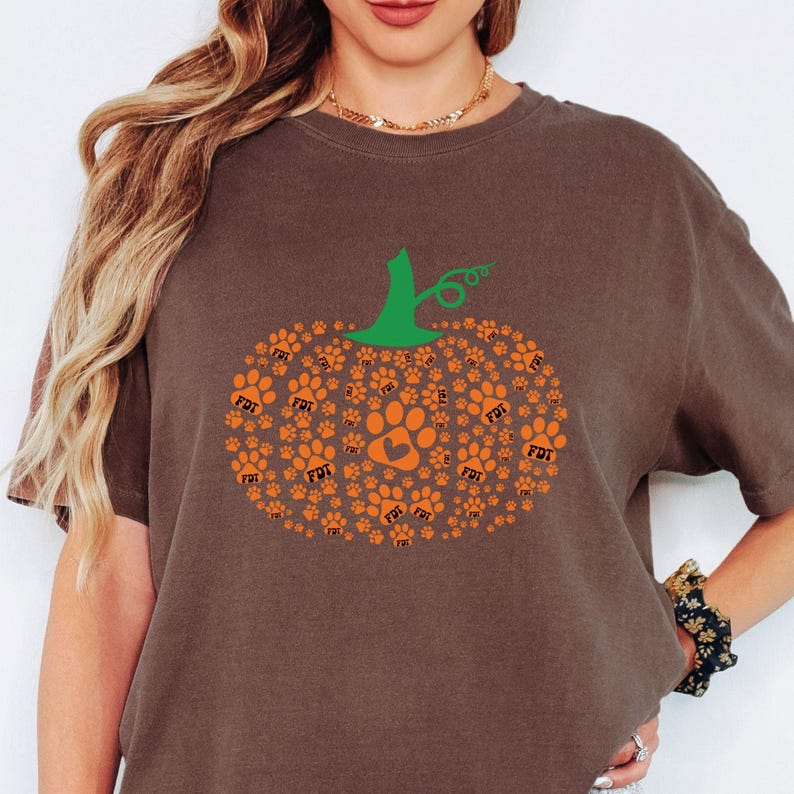 Puede incluir: Camiseta marr&oacute;n con un dise&ntilde;o de calabaza hecho de huellas de patas naranjas y un tallo verde. El dise&ntilde;o de la calabaza incluye una gran huella de pata en el centro. La camiseta tiene las letras "FDT" impresas en algunas de las huellas.