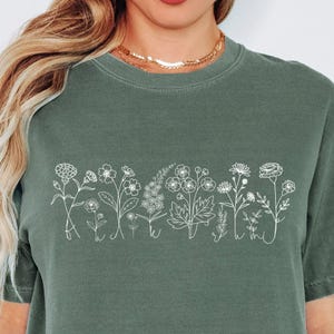 May include: Sage green t-shirt featuring a white line drawing of various wildflowers. The floral design spans the chest area, showcasing detailed botanical illustrations. The shirt has a classic crew neck and short sleeves.