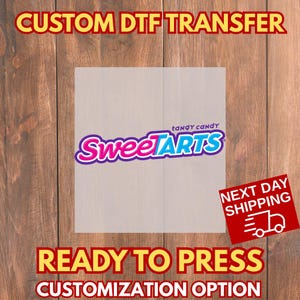 May include: A DTF (Direct to Film) transfer featuring the SweetTarts logo in pink and blue. The words "CUSTOM DTF TRANSFER", "READY TO PRESS", and "CUSTOMIZATION OPTION" are displayed in yellow and red text. The image is on a wooden background.