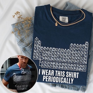 May include: A navy blue short-sleeved t-shirt featuring a white periodic table of elements graphic and the text "I WEAR THIS SHIRT PERIODICALLY" printed in white. The shirt has a Comfort Colors brand label on the inside of the collar.
