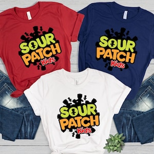 May include: Three t-shirts in red, white, and navy blue, each featuring the "Sour Patch Kids" logo. The logo includes the text "SOUR PATCH Kids" in a mix of green, orange, and red, with a cluster of black candy figures around the text.