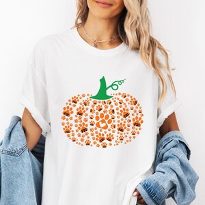 Puede incluir: Camiseta blanca con un dise&ntilde;o de calabaza hecho de huellas de patas naranjas y un tallo verde. Las huellas de las patas tienen las letras "FDT" en su interior. El dise&ntilde;o de la calabaza est&aacute; centrado en la camiseta.