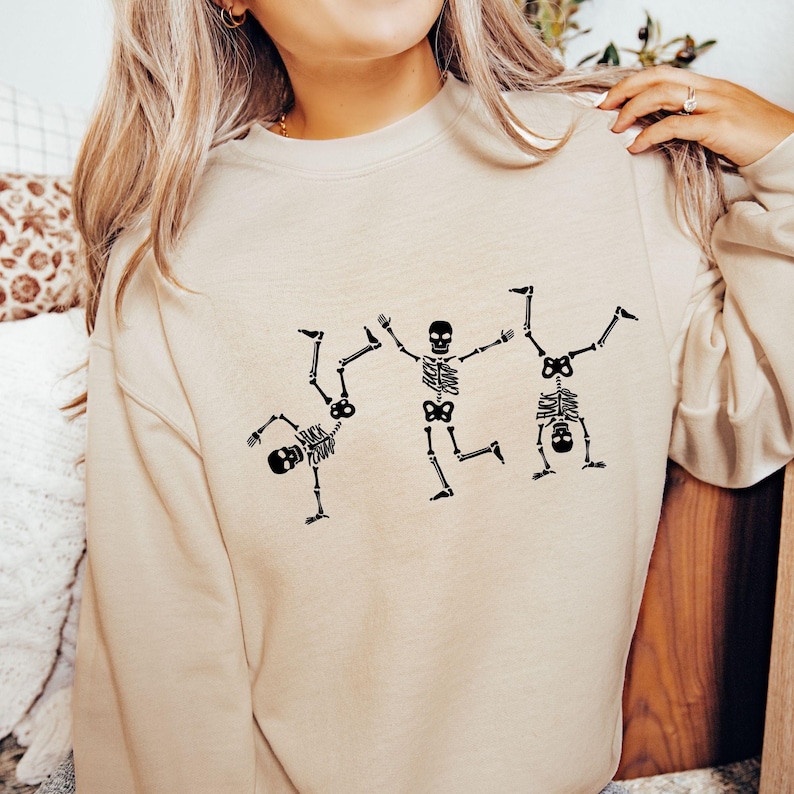 K&ouml;nnte beinhalten: Beiges Sweatshirt mit einem schwarzen Skelett-Design. Die Grafik zeigt drei Skelette in spielerischen Posen, darunter einen Handstand. Ein lustiges und skurriles Design f&uuml;r Halloween oder den Alltag.