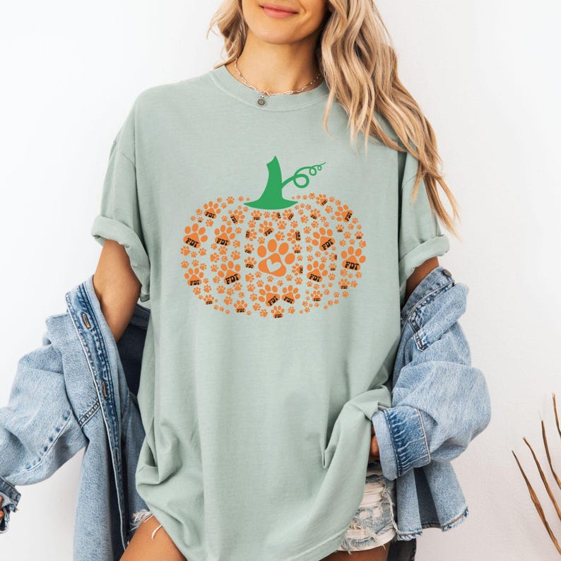 Puede incluir: Camiseta verde claro con un dise&ntilde;o de calabaza hecho de huellas de patas naranjas y las letras "FDT". La calabaza tiene un tallo verde y una huella de pata en el centro. La camiseta es de estilo casual.