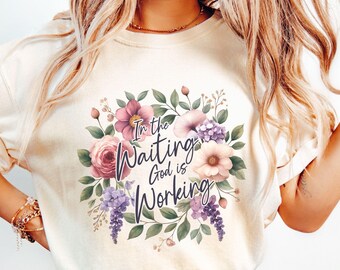 May include: A cream-colored short-sleeve t-shirt featuring a central floral wreath design in shades of pink, purple, and green. The text "In the Waiting God is Working" is printed in a dark blue cursive script in the center of the flowers.