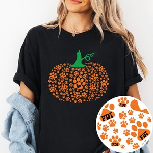 Puede incluir: Camiseta negra con un dise&ntilde;o de calabaza naranja hecho de huellas de patas. La calabaza tiene un tallo verde y las huellas de las patas tienen las letras "FDT" en su interior. Un peque&ntilde;o c&iacute;rculo con huellas de patas est&aacute; en la esquina inferior derecha.