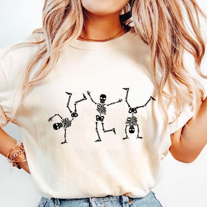 K&ouml;nnte beinhalten: Cremefarbenes T-Shirt mit drei schwarzen Skelettfiguren in spielerischen Posen. Eines der Skelette macht einen Handstand, ein anderes tr&auml;gt den Text "FUCK CAMP" auf dem Brustkorb. Das T-Shirt hat eine lockere Passform.