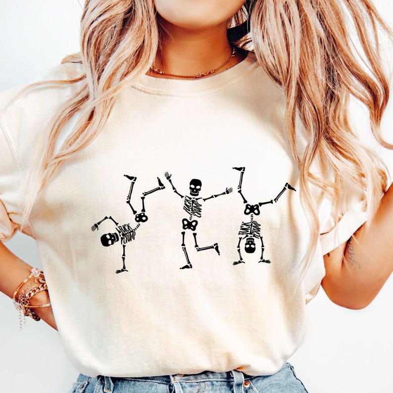 K&ouml;nnte beinhalten: Cremefarbenes T-Shirt mit drei schwarzen Skelettfiguren in spielerischen Posen. Eines der Skelette macht einen Handstand, ein anderes tr&auml;gt den Text "FUCK CAMP" auf dem Brustkorb. Das T-Shirt hat eine lockere Passform.