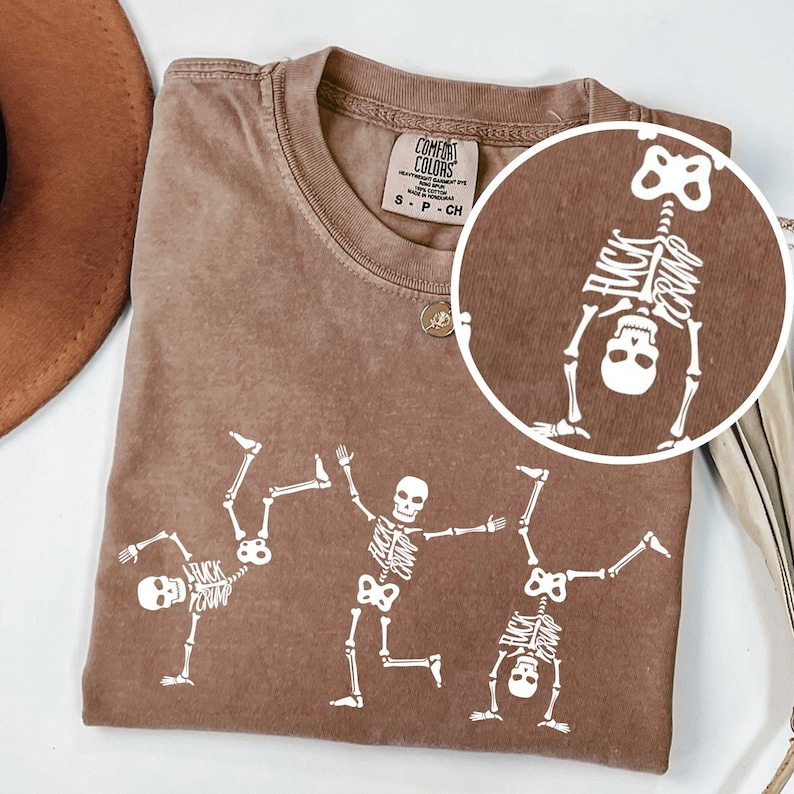 K&ouml;nnte beinhalten: Braunes Comfort Colors T-Shirt mit wei&szlig;en Skelettgrafiken in verschiedenen Posen. Das Shirt enth&auml;lt den Text "FUCK TRUMP" auf den Skeletten. Es hat einen Rundhalsausschnitt und kurze &Auml;rmel.