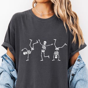 K&ouml;nnte beinhalten: Dunkelgraues T-Shirt mit drei wei&szlig;en Skelettfiguren in spielerischen Posen. Eines macht einen Handstand, ein anderes ein Rad, und das dritte steht mit erhobenen Armen. Das Shirt hat eine lockere Passform und einen Vintage-Look.