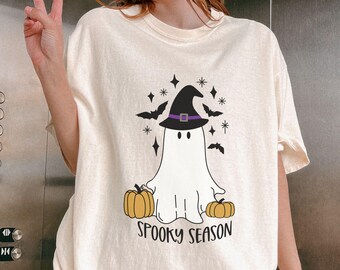 May include: Cream-colored t-shirt featuring a cartoon ghost wearing a witch hat, with pumpkins, bats, and stars. The text "Spooky Season" is printed below the ghost. A fun Halloween-themed graphic tee.