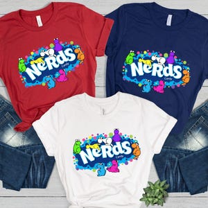 May include: Three t-shirts in red, navy blue, and white, each featuring a colorful "Nerds Candy" graphic. The design includes the word "Nerds" in white, surrounded by cartoon characters in various colors.