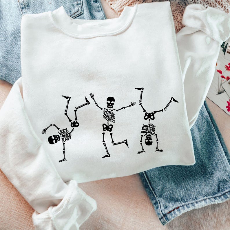 K&ouml;nnte beinhalten: Wei&szlig;es Sweatshirt mit einer schwarzen Grafik von drei tanzenden Skeletten. Ein Skelett macht einen Handstand, ein anderes rennt und das dritte macht einen Purzelbaum. Der Text "FUCK CRAP" befindet sich auf dem Torso des ersten Skeletts.
