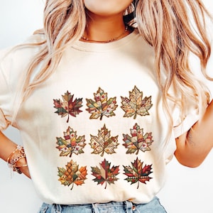 May include: Cream-colored sweatshirt featuring nine colorful maple leaf designs. Each leaf is a patchwork of various patterns and colors, including red, orange, yellow, and green. The leaves are arranged in three rows of three.