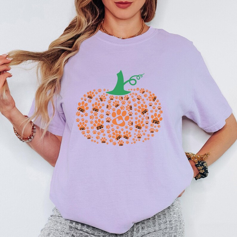 Puede incluir: Una camiseta de color lila claro con un dise&ntilde;o de calabaza hecho de huellas de patas y un coraz&oacute;n de color naranja. La calabaza tiene un tallo verde y est&aacute; centrada en la camiseta. La camiseta es de estilo casual y c&oacute;modo.