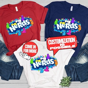 May include: Three t-shirts in red, navy blue, and white, each featuring a graphic of the Nerds candy logo surrounded by colorful cartoon characters. The white shirt includes a red circular graphic with the text Come In For More.