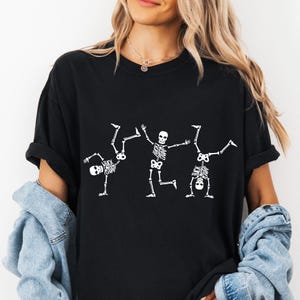 K&ouml;nnte beinhalten: Schwarzes T-Shirt mit einem wei&szlig;en Skelett-Design. Drei Skelette sind in verschiedenen Posen dargestellt. Auf einem Skelett steht der Text "FUCK FUNNY". Das T-Shirt besteht aus einem weichen Material.