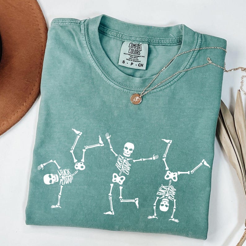K&ouml;nnte beinhalten: Ein t&uuml;rkisfarbenes Comfort Colors T-Shirt mit drei wei&szlig;en Skelettfiguren in spielerischen Posen. Das T-Shirt hat einen Rundhalsausschnitt und kurze &Auml;rmel. Eine silberne Halskette mit Anh&auml;nger liegt auf dem T-Shirt.