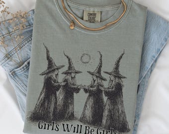 May include: Sage green t-shirt with a graphic of four witches holding hands, with the text "Girls Will Be Girls." A gold necklace rests on the shirt, which is folded on top of a pair of blue jeans.
