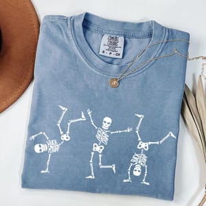 K&ouml;nnte beinhalten: Blaues Comfort Colors T-Shirt mit einem wei&szlig;en Skelett-Design. Drei Skelette sind in spielerischen Posen dargestellt. Eine goldene Halskette mit Anh&auml;nger liegt auf dem Shirt. Der Text "FUCK CRUMP" ist sichtbar. Das Shirt ist in der Gr&ouml;&szlig;e S.