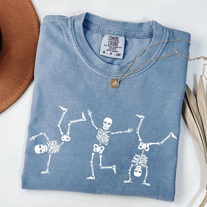 K&ouml;nnte beinhalten: Blaues Comfort Colors T-Shirt mit einem wei&szlig;en Skelett-Design. Drei Skelette sind in spielerischen Posen dargestellt. Eine goldene Halskette mit Anh&auml;nger liegt auf dem Shirt. Der Text "FUCK CRUMP" ist sichtbar. Das Shirt ist in der Gr&ouml;&szlig;e S.