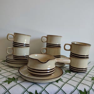 Può includere: Un set da caffè vintage in ceramica color crema con strisce orizzontali marroni. Il set comprende sei tazze, piattini e una lattiera. Le tazze hanno un manico curvo e un bordo leggermente angolato. Il set è disposto su una tovaglia bianca con un motivo floreale verde.