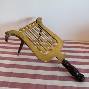 May include: Antique brass lyre-shaped trivet with a dark wooden handle. The trivet has a decorative design with vertical bars and a curved top. The handle is intricately carved and adds to the vintage aesthetic.