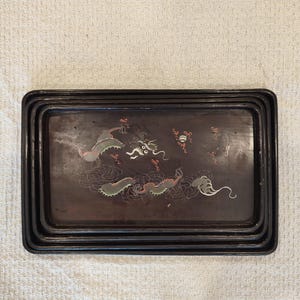 Puede incluir: Un juego de tres bandejas rectangulares anidadas con un acabado marrón oscuro. Las bandejas presentan un diseño decorativo de dragones en verde, blanco y rojo sobre un fondo oscuro. Las bandejas están anidadas.