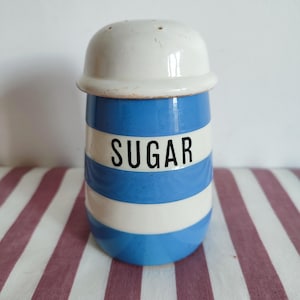 Può includere: Un contenitore per zucchero vintage in ceramica con coperchio bianco e corpo a righe blu e bianche. La parola "SUGAR" è stampata in nero sul davanti. Il contenitore è su una superficie a righe.