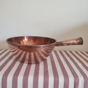 May include: A polished copper bowl with a single long, tapered handle extending from the side. The bowl has a rounded, deep basin and a smooth, reflective metallic surface that shows subtle variations in the warm, reddish-brown copper tone.