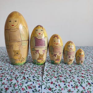 May include: A set of five yellow wooden nesting dolls, each painted with a different girl and a small animal. The dolls are arranged in order of size, with the largest doll on the left and the smallest doll on the right. The dolls are sitting on a blue and white floral tablecloth.