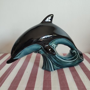 Könnte beinhalten: Eine glänzende Keramik-Delfinfigur mit einem glatten schwarzen Oberkörper, der in einen tiefen blaugrünen Unterteil übergeht. Der Delfin ist in einer Tauchbewegung dargestellt, wobei sich sein Körper über einen stilisierten, wellenförmigen Sockel wölbt.
