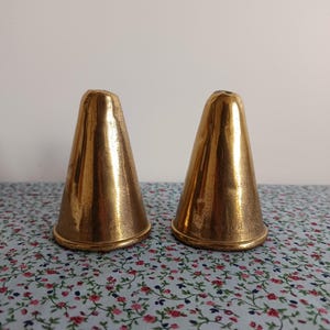 May include: Two antique, gold-coloured, cone-shaped objects. The objects have a textured surface and a rounded base. They are placed on a floral patterned fabric. The objects appear to be decorative or functional items.