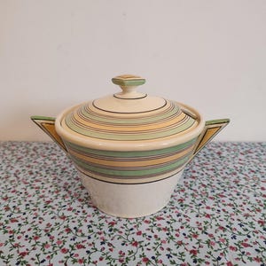 May include: A cream-colored ceramic serving dish with a lid. The lid and rim feature horizontal stripes in yellow, green, and black. The dish has two angular handles and sits on a floral patterned tablecloth.