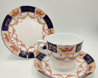 May include: A vintage tea set with a cup, saucer, and plate. The set features a white background with a blue border and orange fruit and floral designs. The cup and saucer are placed on a white surface.