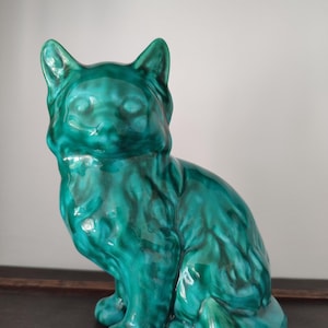 Puede incluir: Figura de gato de cerámica turquesa con acabado brillante. El gato está sentado con las orejas erguidas. La figura tiene una superficie texturizada y el color turquesa tiene acentos verdes más oscuros. Es un artículo decorativo.