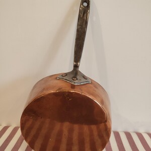 Op de afbeelding: Een koperen steelpan met een lange, geklonken handgreep. De pot heeft een afgeronde bodem en een glanzend oppervlak. De handgreep is gemaakt van donkerder metaal en heeft een gat om op te hangen. De pan wordt waarschijnlijk gebruikt om te koken.