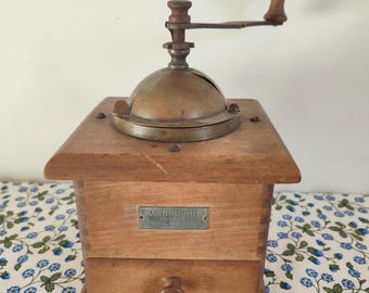 Op de afbeelding: Vintage houten koffiemolen met een messing bovenkant en hendel. De molen heeft een lade en een klein metalen plaatje met tekst. De houten behuizing is warmbruin van kleur en de hendel is van hout.