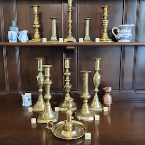 May include: A collection of brass candlesticks of varying heights and designs, alongside small ceramic pitchers and a glass bottle, each labelled with a letter from A to K, arranged on two wooden shelves with a dark wood panel background.