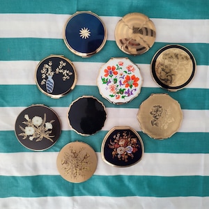 May include: A collection of vintage compact mirrors with various designs. The compacts feature gold, black, and navy blue cases with floral, geometric, and starburst patterns. Some have scalloped edges, and others are round or octagonal.