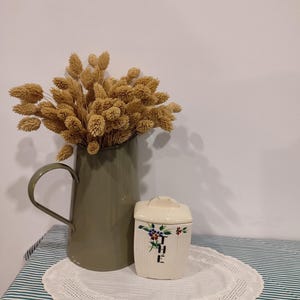 May include: A green metal jug with dried flowers and a white ceramic canister with a floral design and the word "THE" on the front.